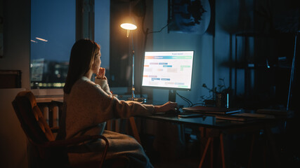 Female Manager Works on a Desktop Computer in Creative Agency in Dark Loft Office in the Evening. Renovated Stylish Design with House Plants, Posters and Rounded Windows. Over the Shoulder Shot.