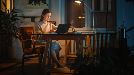 Young Successful Female Works on a Laptop Computer in Creative Agency in Loft Office in the Evening. Renovated Stylish Design with House Plants, Artistic Posters and Big Windows.