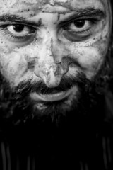 Tight black and white portrait of a bearded middle eastern man with the face covered by mud and an intense gaze	