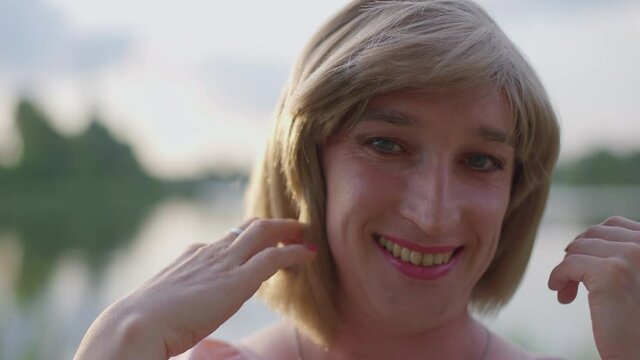 Headshot of smiling trans woman touching hair looking at camera at sunset outdoors. Close-up portrait of joyful carefree Caucasian non-binary person resting at lake on summer evening