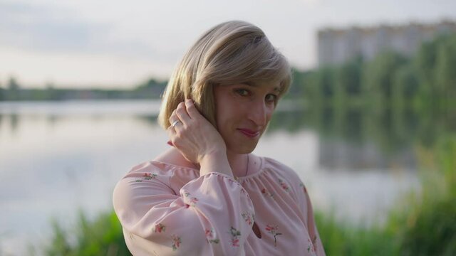 Portrait of shy Caucasian transgender woman posing outdoors at sunset on lake. Timid non-binary gender-fluid person in pink dress looking at camera smiling. Lifestyle and gender identity