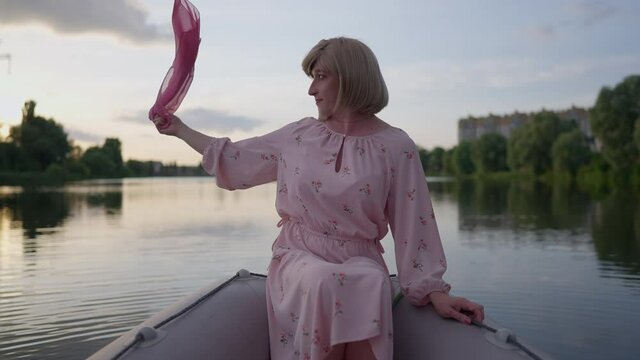 Happy carefree non-binary person admiring sunset on lake shaking pink kerchief in hand. Portrait of relaxed satisfied Caucasian transgender woman in romantic dress dating outdoors at dusk