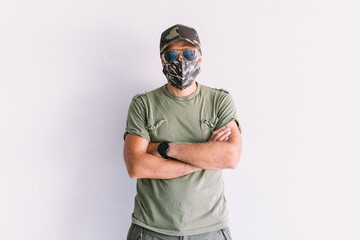 Military man with camouflage cap, sunglasses and camouflage mask, with crossed arms, on a white wall