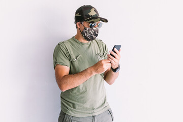 Military man wearing camouflage cap and sunglasses, looking at mobile phone, on a white wall