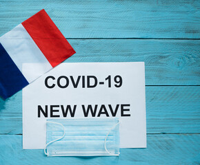 Fototapeta na wymiar On a blue wooden background is a sheet with the inscription: covid -19 new wave, a medical mask and the flag of France