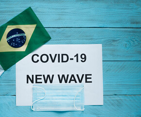 Fototapeta na wymiar On a blue wooden background lay a sheet with the inscription: covid -19 new wave, a medical mask and the flag of Brazil