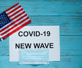 Fototapeta na wymiar On a blue wooden background is a sheet with the inscription: covid -19 new wave, a medical mask and a U.S. flag