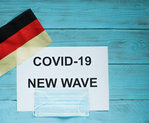 Fototapeta na wymiar On a blue wooden background is a sheet with the inscription: covid -19 new wave, a medical mask and a German flag