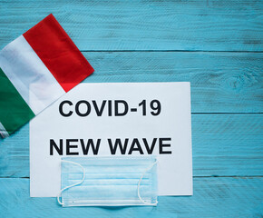 Fototapeta na wymiar On a blue wooden background are a sheet with the inscription: covid -19 new wave, a medical mask and the flag of Italy