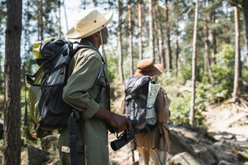 African american hiker with backpack and binoculars walking near blurred wife in forest.