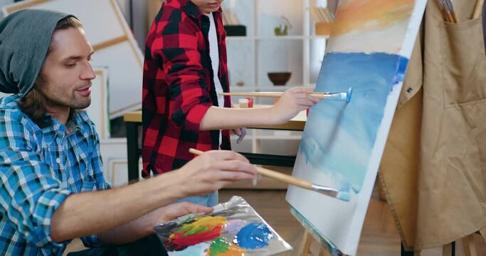 Attractive positive creative modern male artist and cute boy drawing blue sea on joint future picture attached to easel in home studio