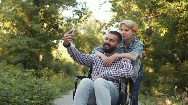 Woman hugs man in wheelchair who is taking selfie. Happy couple smiles and takes photos while walking in park. Concept of supporting people with disabilities