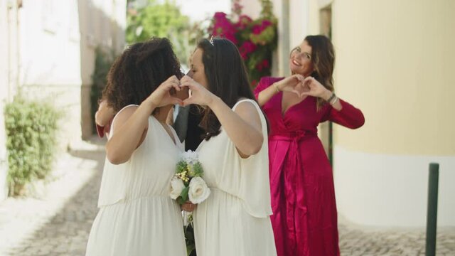 Brides having photoshoot with friends after wedding ceremony. Afro-American lesbian couple making heart with their hands, kissing and posing for camera on summer day. Love, LGBT wedding concept