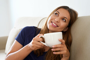 Pretty positive dreamy girl holds cup with warm beverage imagining sitting on sofa