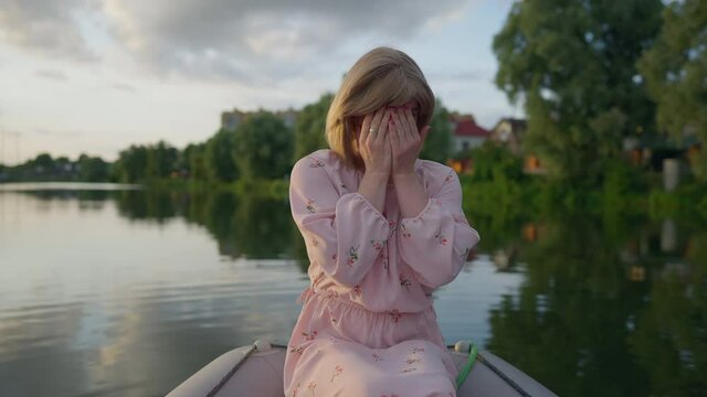 Modest Caucasian transgender woman laughing closing face with hands looking at camera. Positive queer non-binary person sitting in boat on summer lake having fun. Shyness and timidity concept