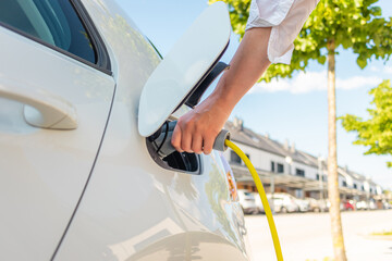 Woman plugging in the charger into a socket of her white electric car at a charging station in the...