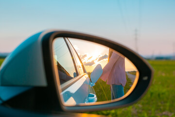 Fototapeta premium Rear mirror with a reflection of the female silhouette charging an electric car, in the background visible sky at sunset