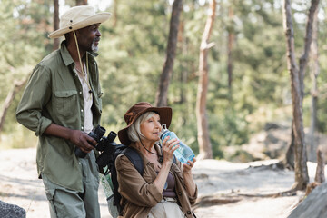 African american tourist holding binoculars near senior wife with water in forest.
