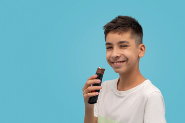 Waist up portrait of a young smiling boy teen eating chocolate against  blue background in studio