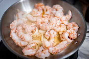 Macro closeup of stainless steel frying pan with sauteing olive oil, sweet onion slices and whole large king jumbo Argentinian shrimp seafood cooked on electric stove in home kitchen