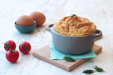 Close up of French souffle omelette in mini casserole with cherry tomatoes, eggs and herbs on a wooden board in background 