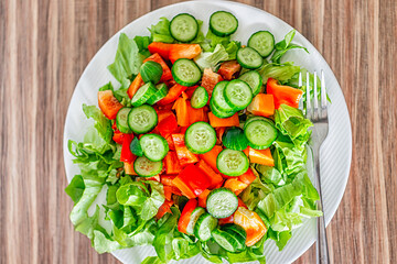 Closeup flat top macro of fresh raw chopped vegetable salad with romaine lettuce greens, sweet pepper, cucumbers on white plate, healthy lunch meal by fork on wooden table