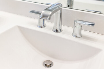 New modern stainless steel metal faucet with handles in bathroom room white porcelain sink closeup by mirror with drain hole