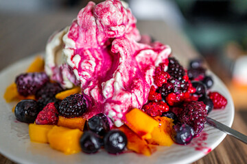 Macro closeup of colorful bowl white plate of vanilla banana vegan ice cream topped with frozen blueberries, red raspberries berries and mango with spoon on wooden kitchen table