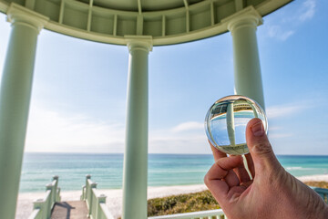 Seaside, Florida with man hand holding lensball crystal glass ball with reflection of green wooden pavilion gazebo architecture by beach ocean in background view on sunny day - obrazy, fototapety, plakaty