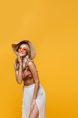 joyful woman in ruffle bikini top, white skirt and sun hat isolated on yellow.