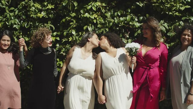 Slider shot of lesbian brides taking photo with friends. Cheerful women in evening wears standing in line, holding hands and posing for camera. LGBT wedding, photoshoot concept