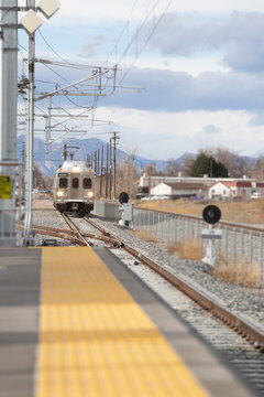 Train arriving at the station. Lights on coming down the tracks. Pedestrian platform in foreground. Rocky Mountains in background.