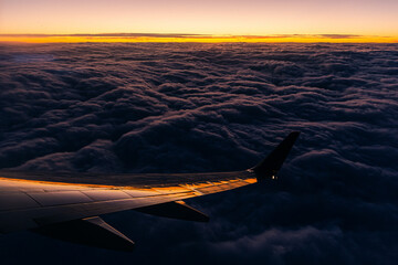 Sunset raking over the top of clouds. View from an airplane window at dusk. Birds eye view with plane wing in foreground.
