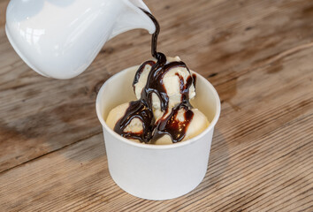 Three scoops of white ice cream with chocolate filling in a cardboard cup on a rough wooden table.