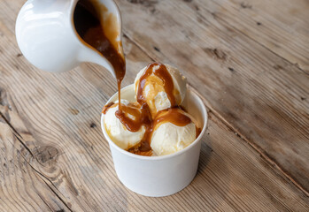 Three scoops of white ice cream are poured over caramel syrup in a cardboard cup on a rough wooden table.