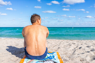 Turquoise vibrant colorful water and back of tan man sitting on towel looking at sand shore in...