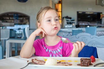 cute four years old blonde girl eating with spoon sweet dessert: black chocolate, vanilla ice cream, strawberries and mango sauce, sitting in restaurant
