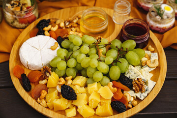 cheeses with grapes, dried fruits, nuts, honey and jam on a round wooden plate.