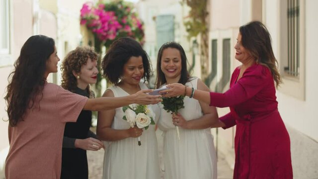 Medium shot of lesbian brides taking photo with friends. Beautiful women in evening wears celebrating wedding, taking selfie from different angles, posing for camera. Photoshoot, LGBT wedding concept