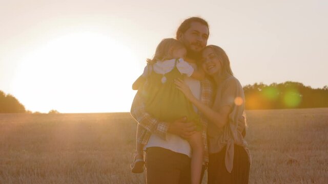 Happy group of people caucasian family young parents with small child woman and man holding daughter in arms little baby girl stand hugging cuddling standing hug in wheat field enjoy sunbeams sunset