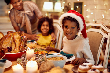 Happy black boy wears Santa cap and enjoys in Christmas dinner with his family at dining table.