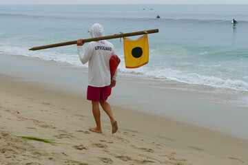 lifeguard walking on the beach carrying floatation device and flag on pole - Powered by Adobe