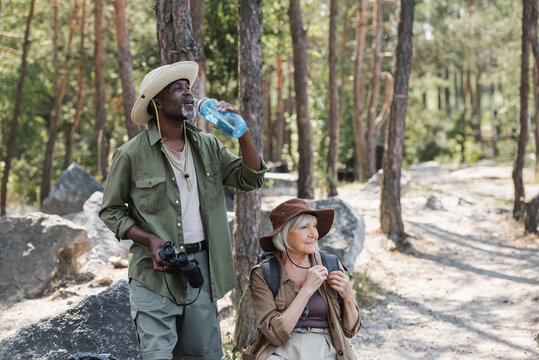 African american traveler holding bottle of water and binoculars near smiling wife in forest. - Powered by Adobe