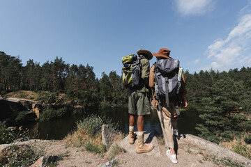 Fototapeta na wymiar Back view of multiethnic hikers with backpacks standing on rock near lake and forest.