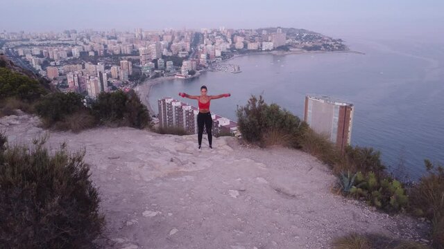 Video 4K, de chica haciendo ejercicio de hombros con mancuernas en la monta&ntilde;a, con vistas al mar y a la ciudad.