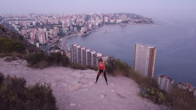 Video 4K, de chica haciendo sentadillas con mancuernas, en lo alto de la monta&ntilde;a con vistas al mar y a la ciudad.