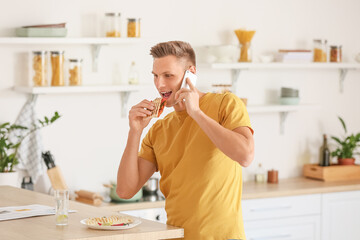 Handsome young man talking by phone while eating tasty quesadilla at home