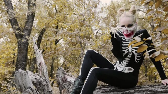 A girl with a scary make-up in a skeleton costume, a mask with rhinestones in a gloomy autumn forest. Sits on a withered old tree. waving bones instead of hand. Halloween 