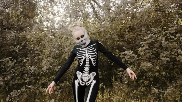 A girl with a scary make-up in a skeleton costume, a mask with rhinestones in a gloomy autumn forest. Goes to the camera, zombie movement. Halloween, autumn holiday concept. Slow motion, 4k 