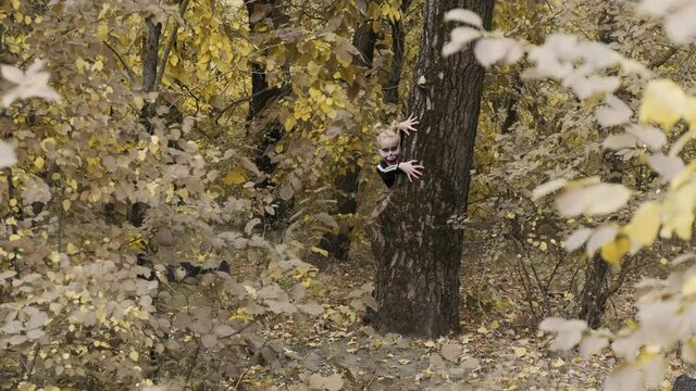 A girl with a scary make-up in a skeleton costume, a mask with rhinestones in a gloomy autumn forest. Climbs out from behind the tree. Halloween, autumn holiday concept. Slow motion, 4K footage